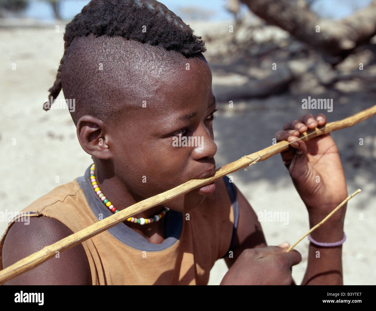 La Namibia, il Kaokoland, Epupa. Un Himba della gioventù, il suo stile di capelli in una lunga treccia chiamato ondatu, riproduce una bocca bow - uno strumento musicale favorito da ragazzi. Esso è costituito dal Makalani palm.Gli Himba sono Herero-parlando Bantu nomadi che vivono nel duro, secco ma fortemente bellissimo paesaggio del nord-ovest remota Namibia. Foto Stock