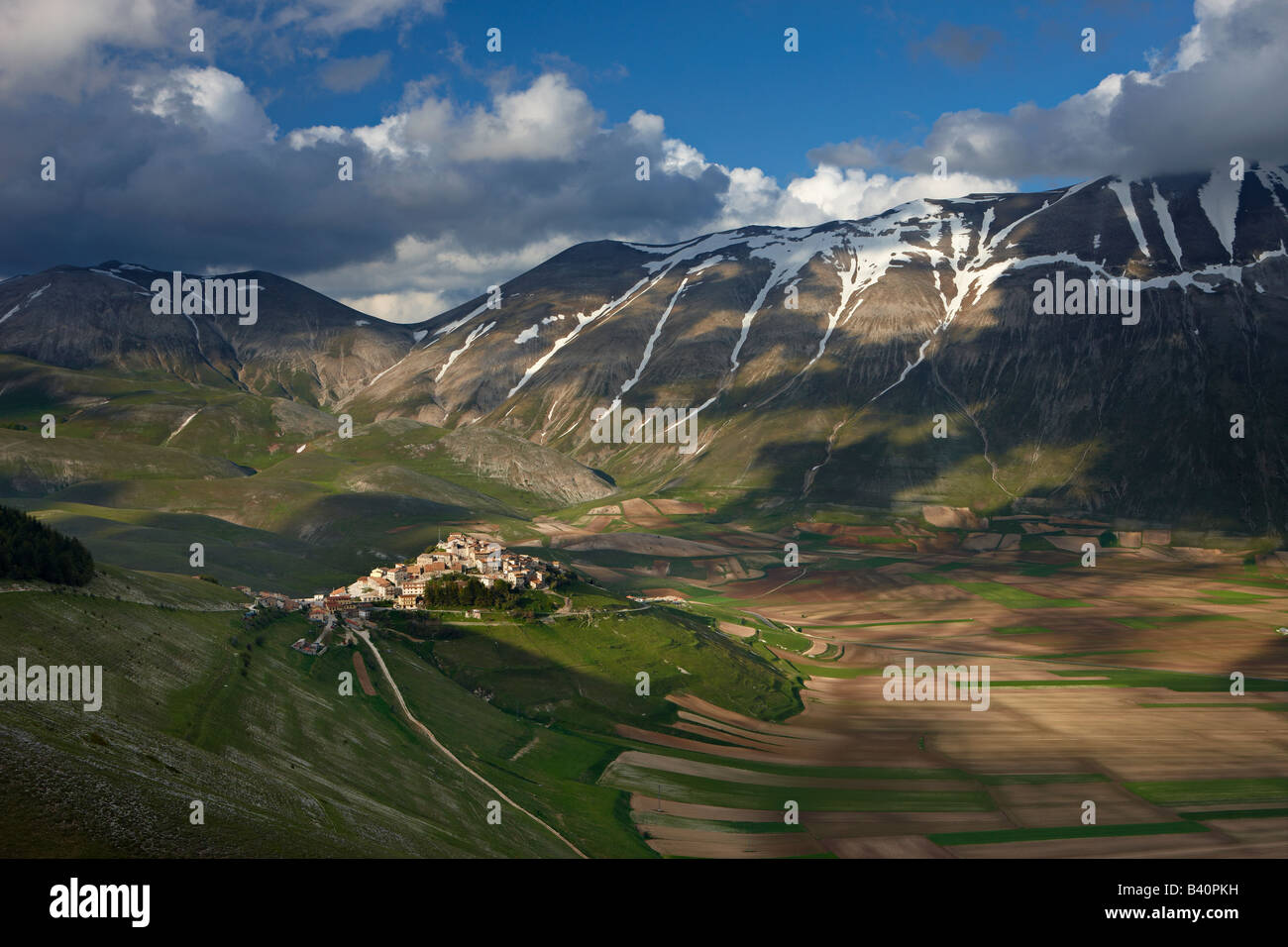 Il borgo di Castelluccio arroccato sopra il piano Grande con le montagne del Parco Nazionale dei Monti Sibillini, Umbria, Italia Foto Stock