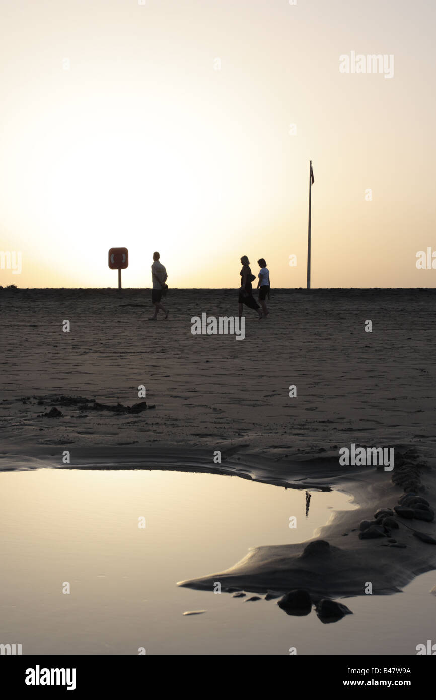 Una famiglia di tre stagliano dal sole al tramonto a piedi passato una bandiera rossa e lifebouy e una piscina di acqua di mare al tramonto sulla spiaggia Foto Stock