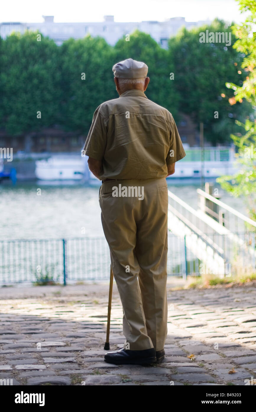 Un uomo anziano si sofferma a contemplare un fiume mentre si cammina in un parco nel tardo pomeriggio aiutata da un bastone da passeggio. Foto Stock