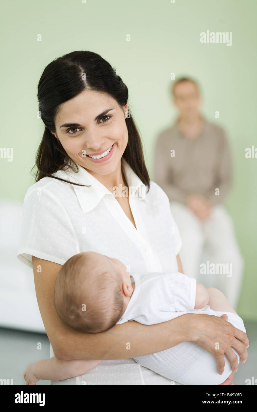 Azienda madre baby, sorridente alla fotocamera, uomo in background Foto Stock