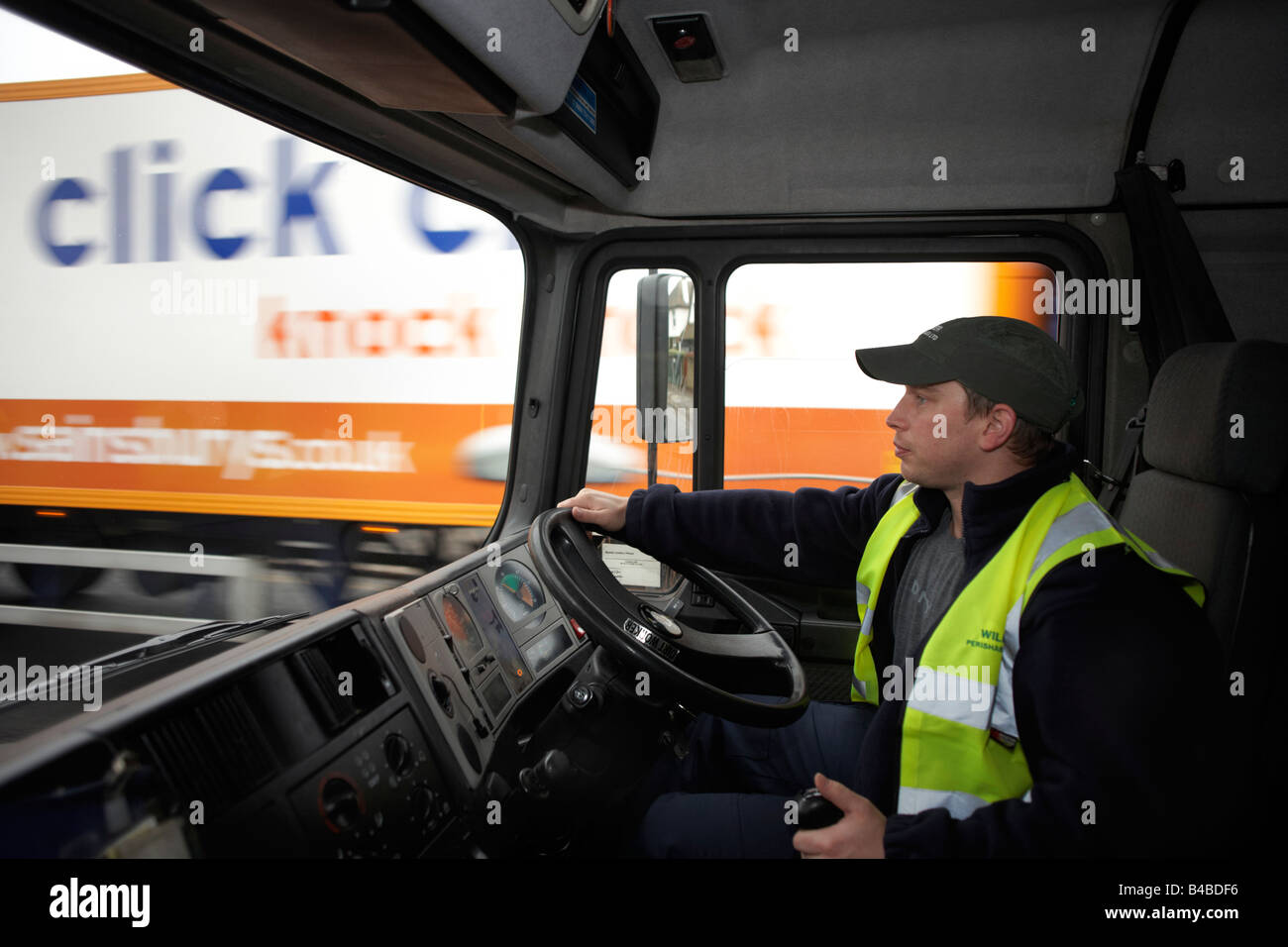 Indossando un alto-vis tabard e berretto, un giovane conducente siede al volante del suo camion dei mezzi pesanti sulla A3 trunk Road a Londra Foto Stock