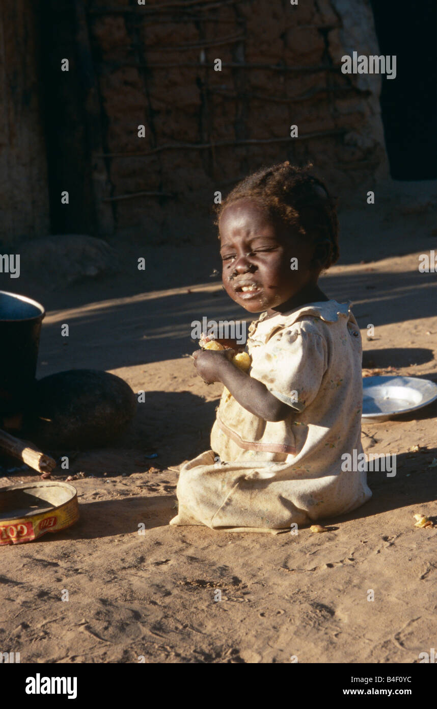 Bambino mangiare sul suolo strizzare gli occhi dai riflessi del sole a sfollati camp, Angola Foto Stock