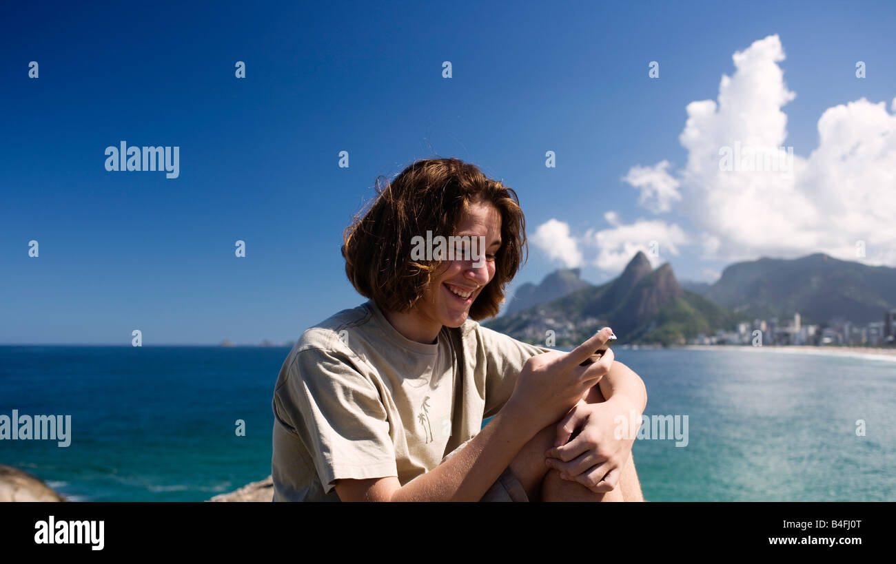 Adolescente 14 a 17 anni di età la lettura di un messaggio di testo sul telefono cel con la spiaggia di Ipanema in background Foto Stock