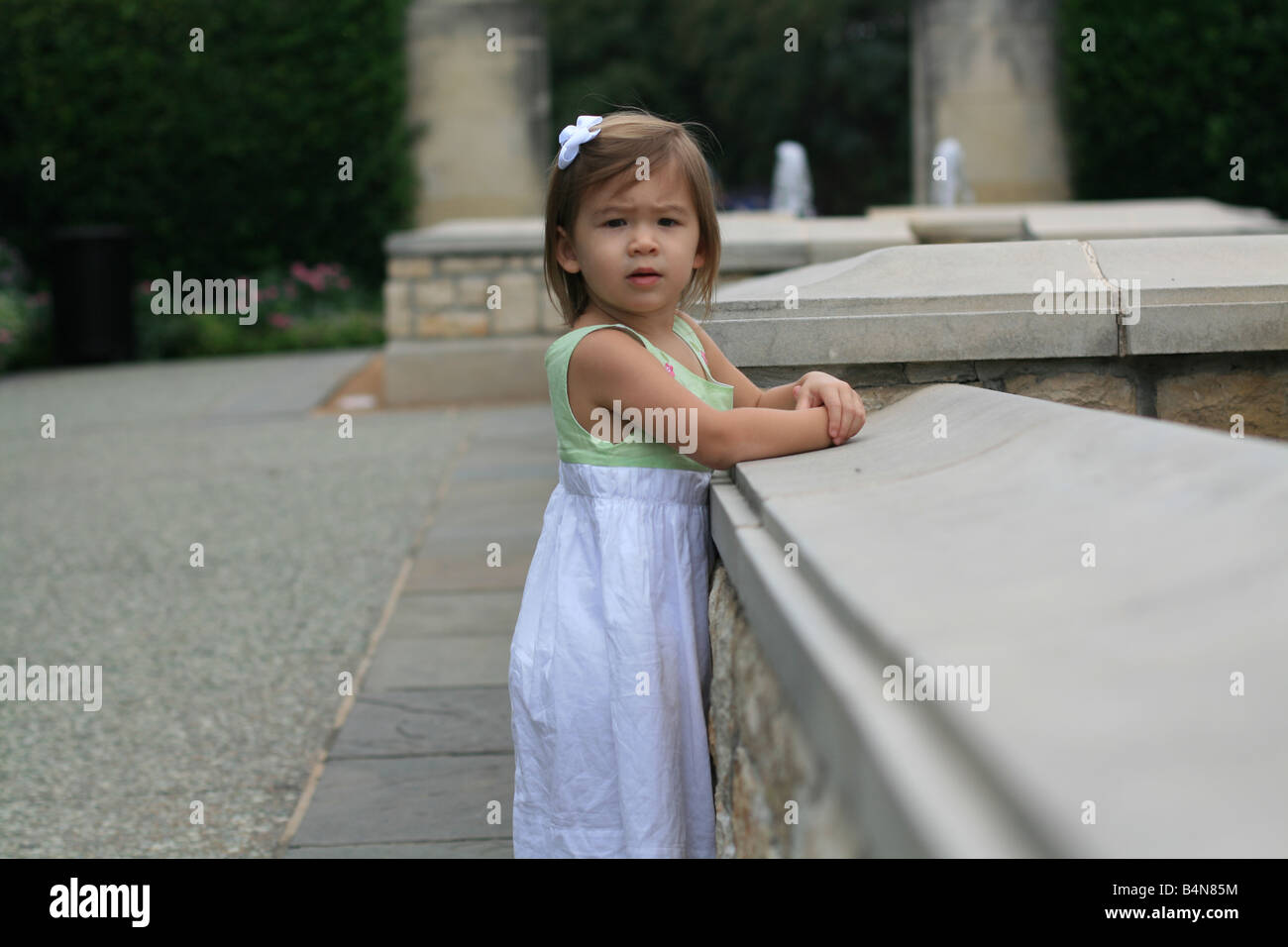 Un due-anno-vecchia ragazza guarda alla sua destra mentre in piedi da una fontana. Foto Stock