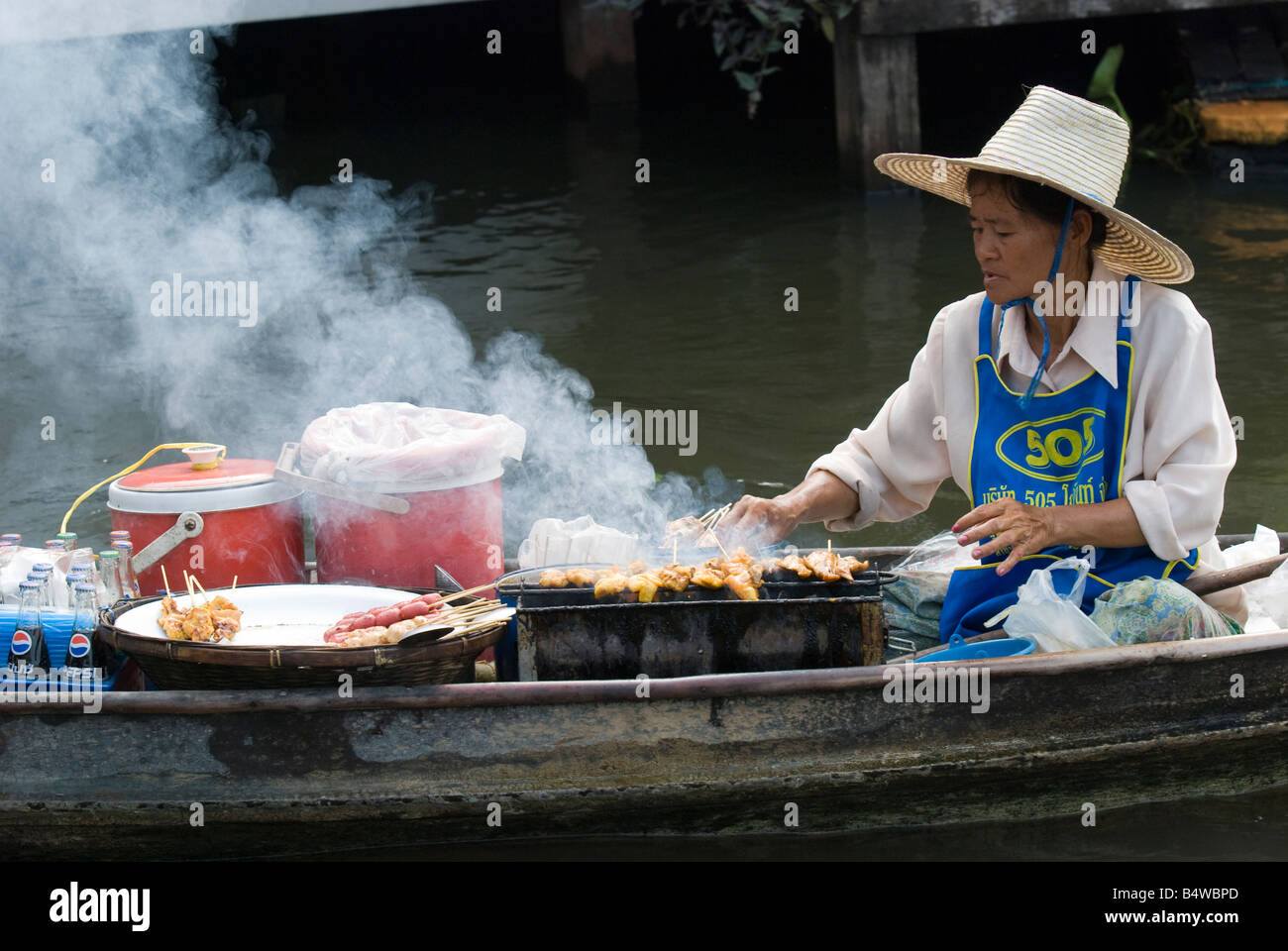 Donna anziana cuocere carne su un piccolo barbecue in una barca di legno tradizionale su un canale in Thailandia Foto Stock