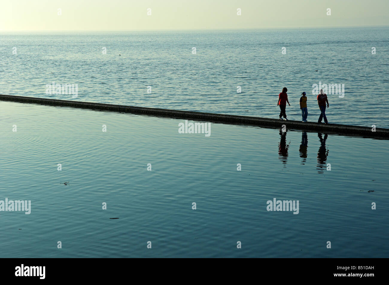 Tre persone camminando lungo la parete della piscina infinity affacciata sul mare di sera presto Foto Stock