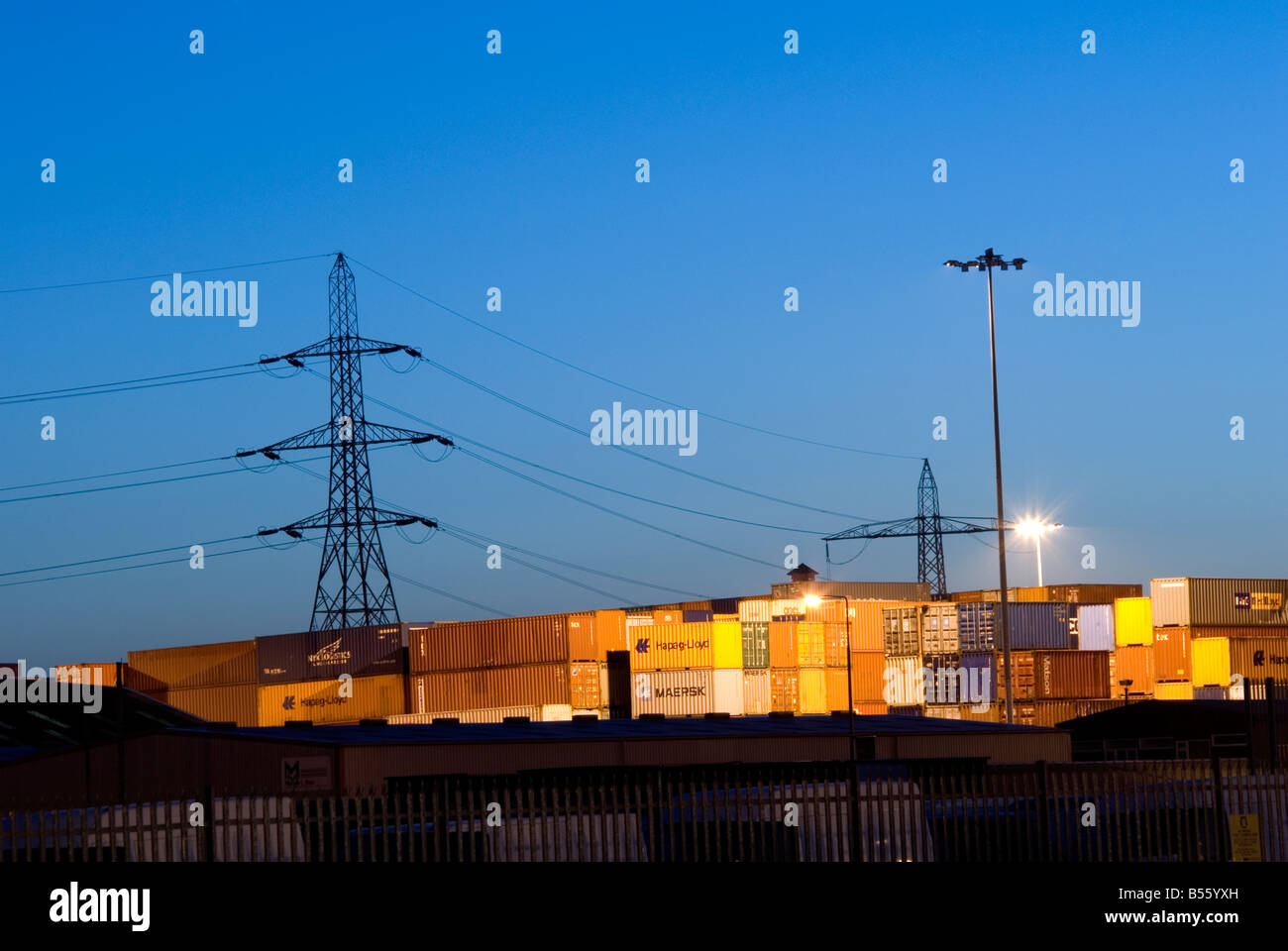 Una vista di grandi contenitori di deposito presso sunrise illuminato da luci spot con elettricità tralicci stagliano in background Foto Stock