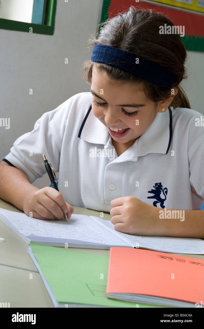 Junior schoolgirl felicemente si concentra sul suo iscritto alla sua scrivania in aula scolastica Foto Stock