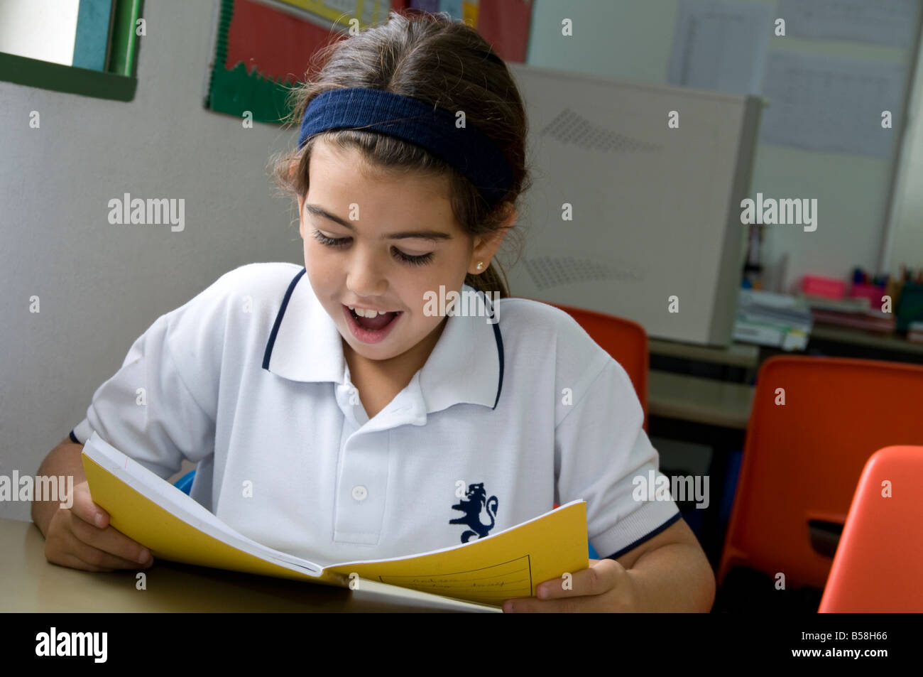 Junior schoolgirl felicemente si concentra su di lei la lettura alla sua scrivania in aula scolastica Foto Stock