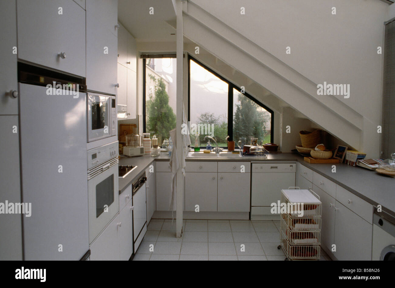 Moderna cucina bianca sotto le scale con soffitto inclinato e finestra con vista sul cortile giardino Foto Stock
