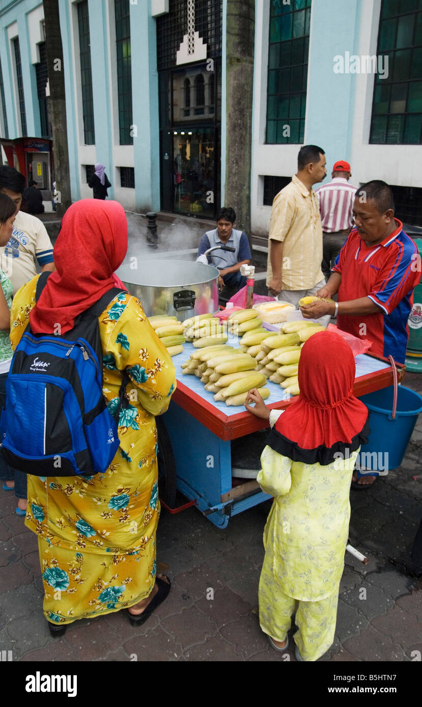 Una donna e bambino indossa red head sciarpe ordinare alcuni hot sulla pannocchia di mais da un venditore ambulante di Kuala Lumpur in Malesia Foto Stock
