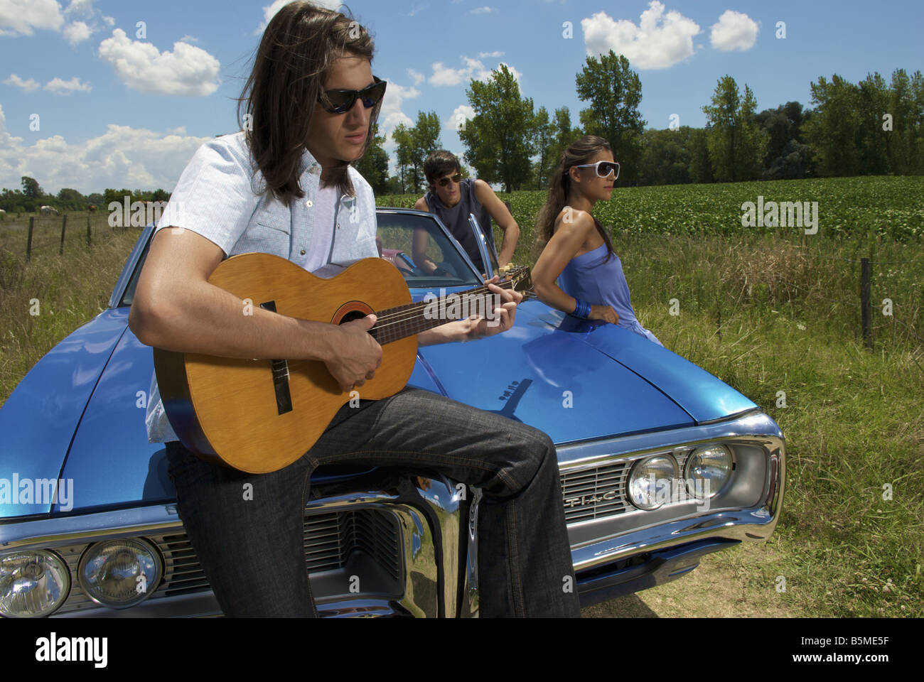 Tre giovani adulti con un auto blu a suonare la chitarra nel paese Foto Stock