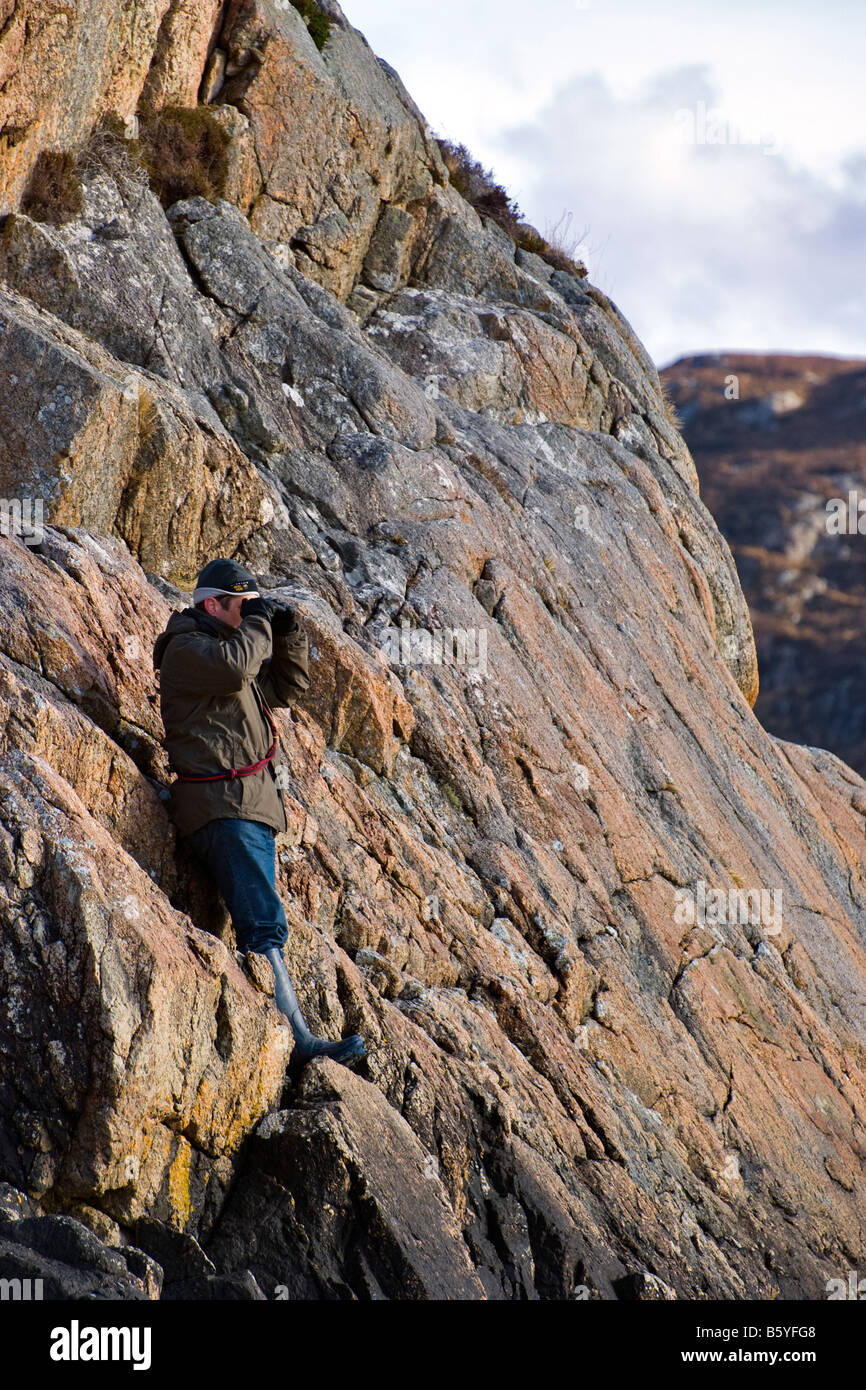 Uomo in abbigliamento da esterno in piedi a metà della scogliera rocciosa, guardando verso il mare con le mani che proteggono gli occhi dal sole, Wester Ross, Scozia Foto Stock