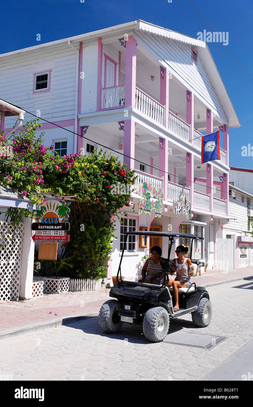 Due donne in un golf cart passato alla guida di una casa in legno dipinto in colori Caraibici, San Pedro, Ambergris Cay Isola, Belize, Cento Foto Stock