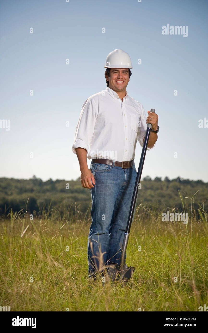 Giovane uomo ispanico in un campo con una pala e indossa un elmetto e sorridente alla fotocamera. Foto Stock