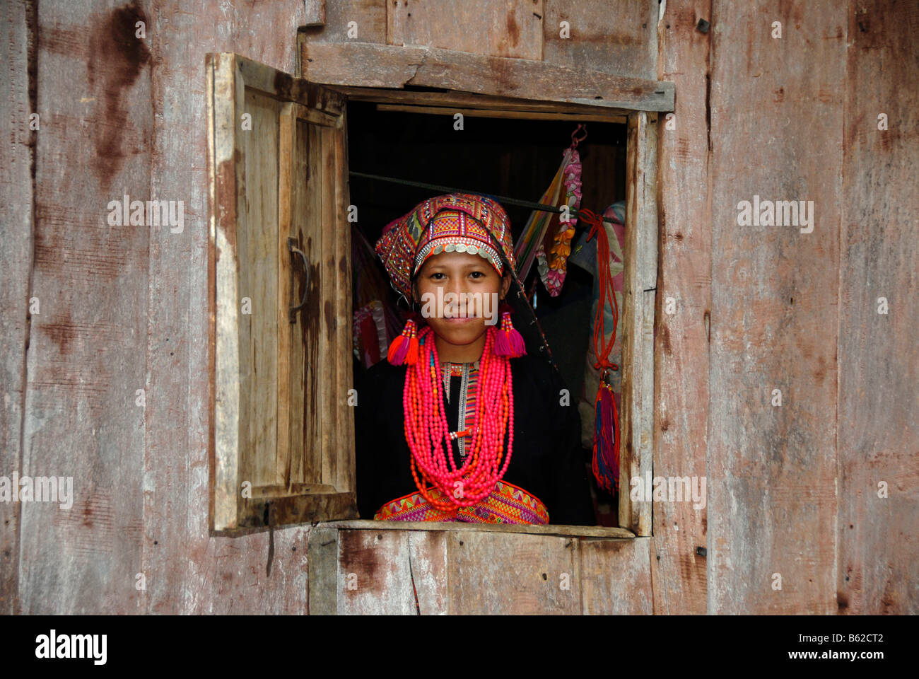 Giovane donna di Akha Pala tribù guardando fuori della finestra un vestito in un colorato testa-vestito di rosso e un necklace, Ban Saenkham T Foto Stock
