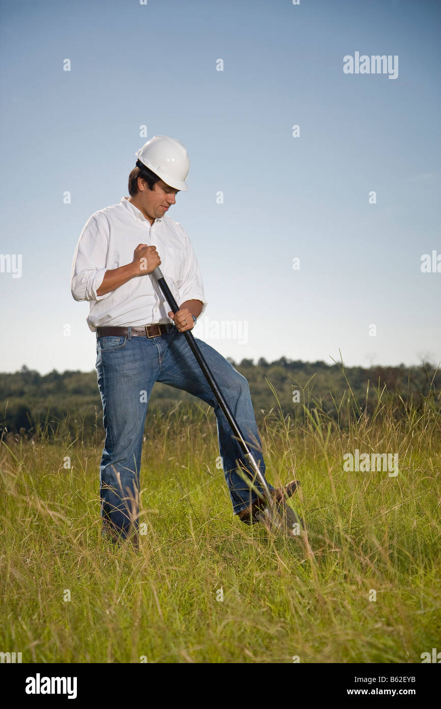 Uomo ispanico in un campo che indossa un elmetto e lo scavo nel terreno con una pala. Foto Stock