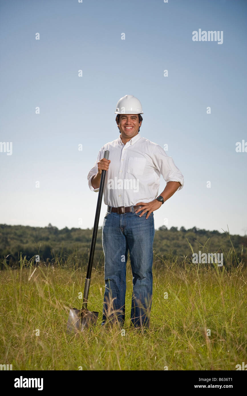 Giovane uomo ispanico in un campo con una pala e indossa un elmetto e sorridente alla fotocamera. Foto Stock