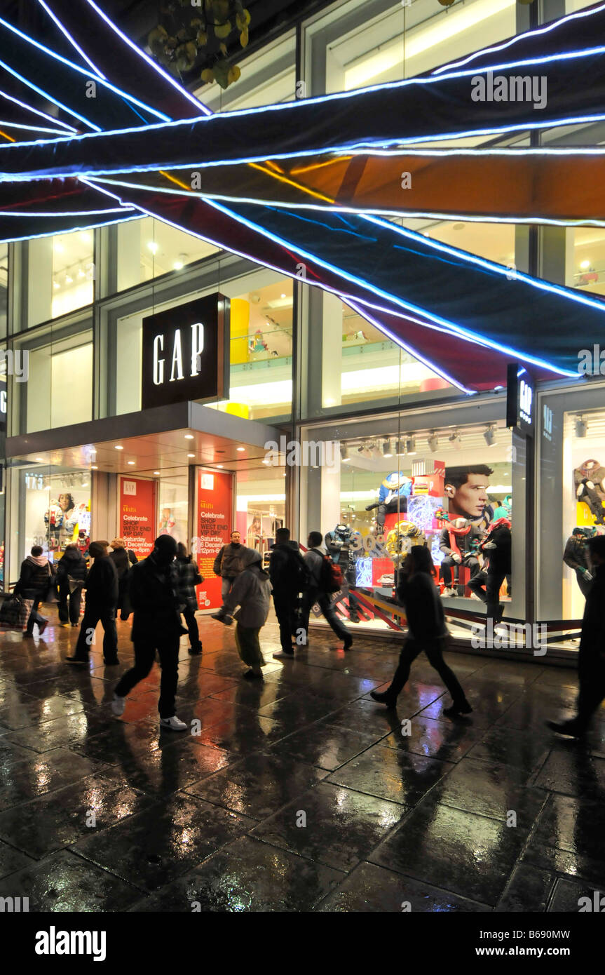 I clienti bagnati fuori dal negozio di moda Gap e dal negozio Window Oxford Street London West End di notte gente Natale Shopping nella pioggia Inghilterra Regno Unito Foto Stock