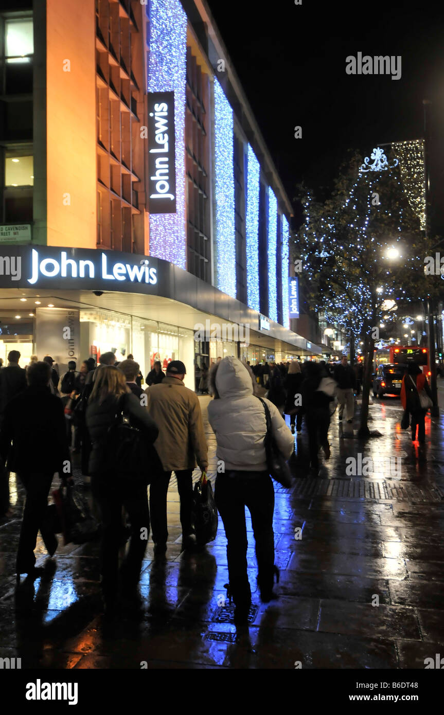 Gli acquirenti che camminano su marciapiedi bagnati John Lewis grande magazzino Oxford Street a Londra West End con luci di strada di Natale e decorazioni Inghilterra Regno Unito Foto Stock