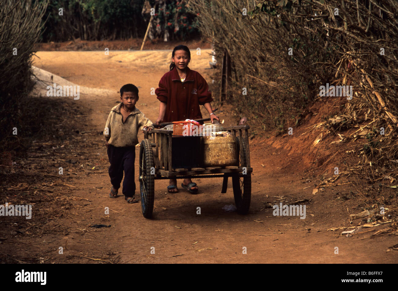 Una ragazza Hmong e suo fratello a spingere un carrello a mano lungo la corsia polveroso di un villaggio hmong vicino a Phonsavan, Xieng Khuang Prov. Laos Foto Stock
