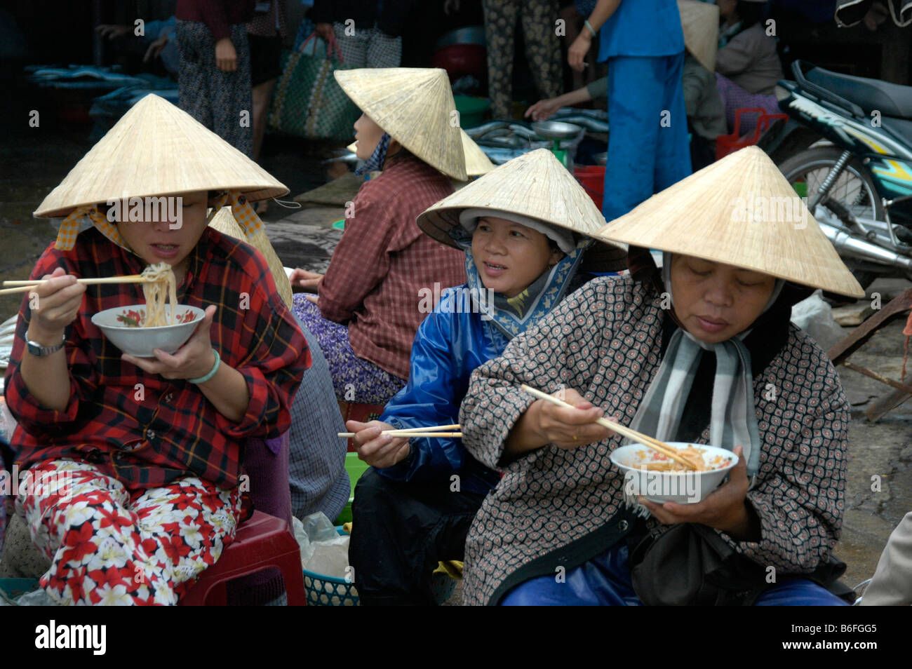 Donne che indossano conformata a cono cappelli di mangiare la zuppa di noodle con i bastoncini presso il mercato del pesce, Hoi An, Vietnam, sud-est asiatico Foto Stock