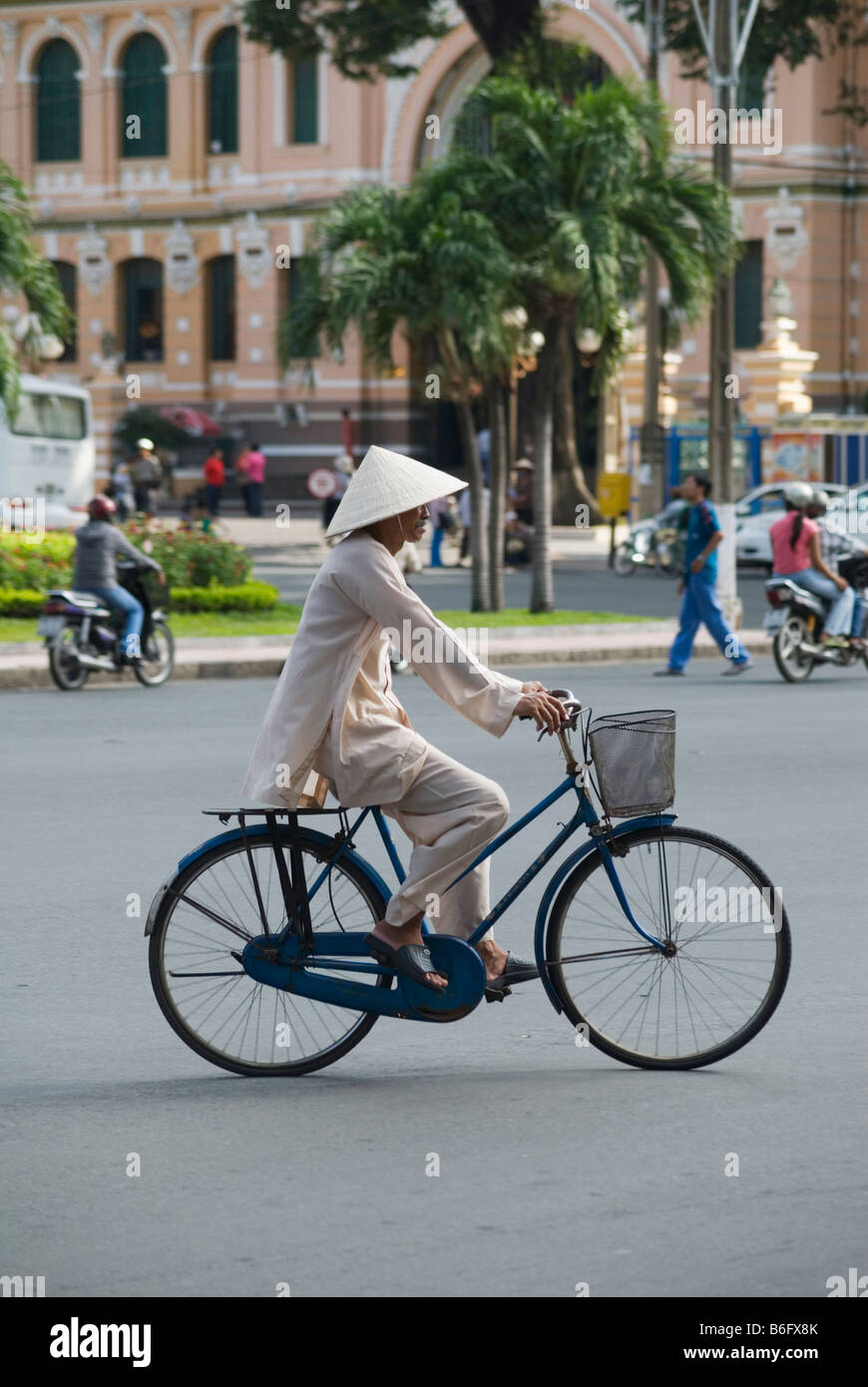Un uomo che indossa tradizionale vietnamita vestiti e cappello conico, escursioni in bicicletta lungo una strada nel centro di Ho Chi Minh City, Vietnam Foto Stock