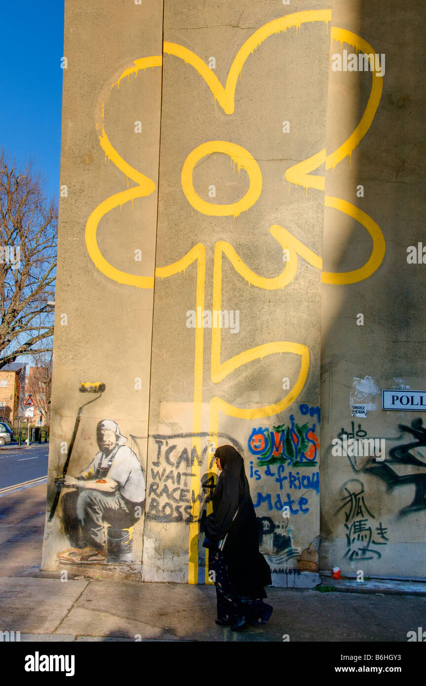 Una donna musulmana oltrepassando Banksy la linea gialla fiore graffiti, Pollard Street, Bethnal Green, Londra,UK Foto Stock