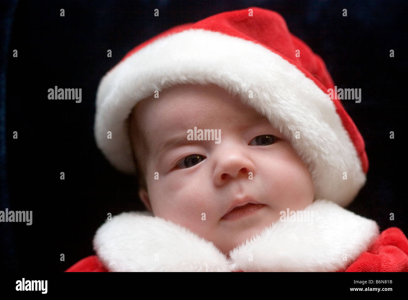 Tre mesi di età bambina vestito in un abito di Santa e cappello di Natale per contro uno sfondo scuro Foto Stock