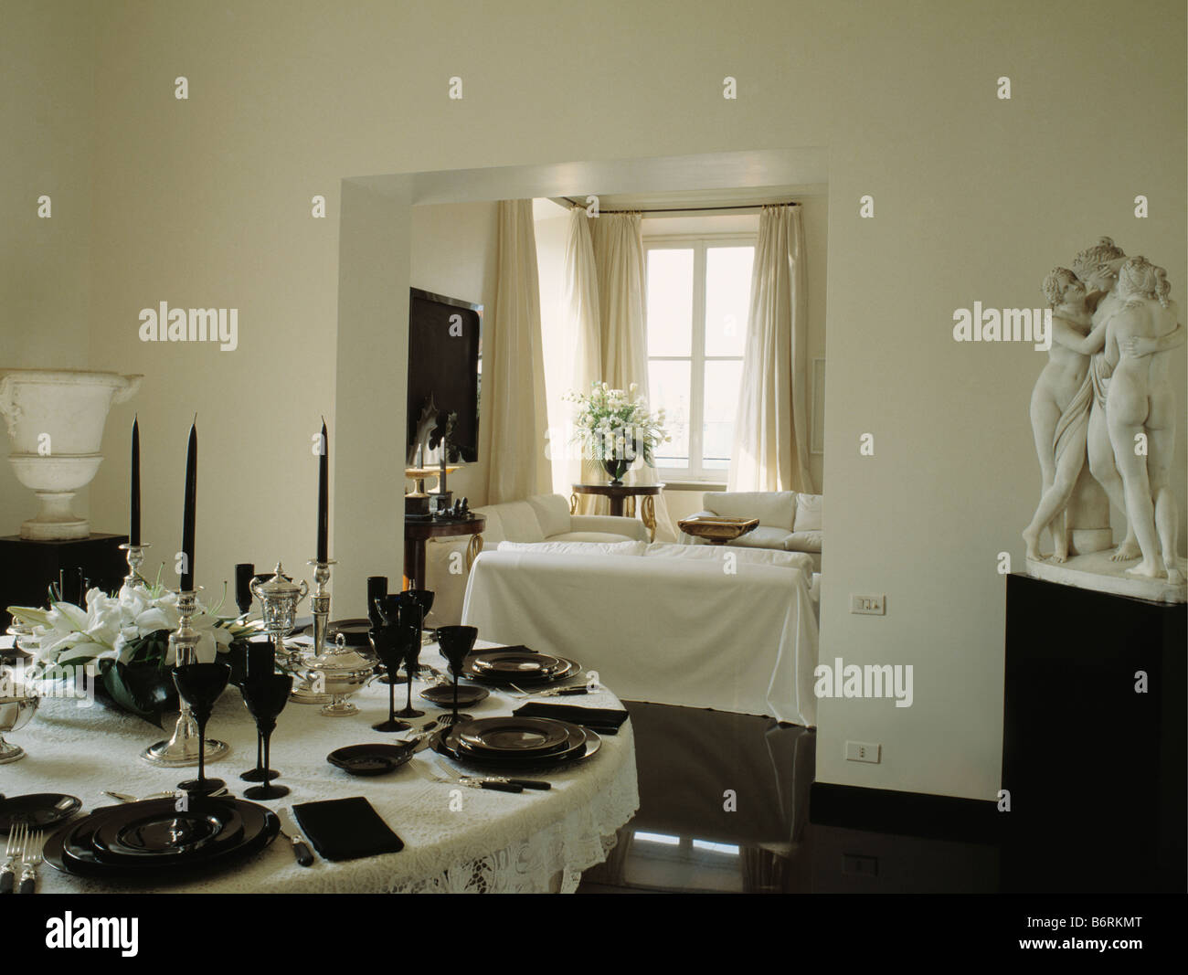 Vasellame nero e candele sulla tavola circolare con un panno bianco in tutto bianco sala da pranzo con statua classica in un angolo della stanza Foto Stock