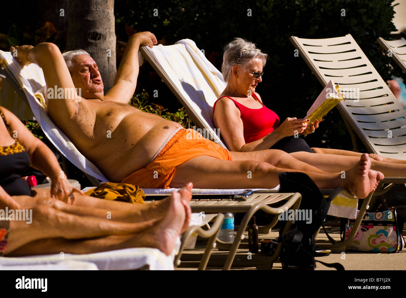 Palm Beach Shores , coppia di grandi di mezza età dai capelli grigi uomo su una sedia a sdraio in arancione costume da bagno sunbathes dalla piscina Foto Stock