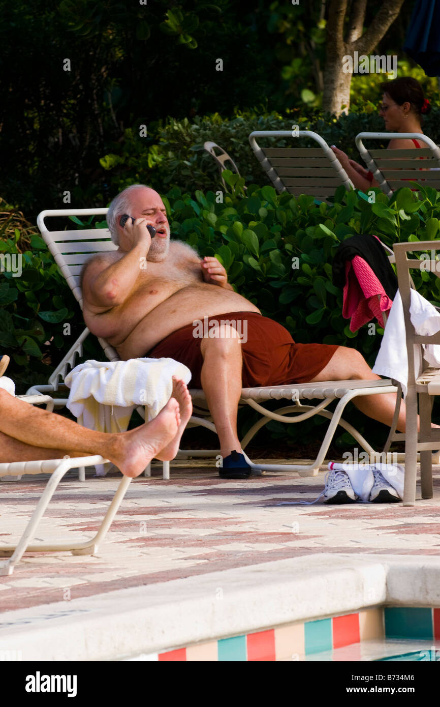 Palm Beach Shores , piscina , dai capelli grigi grande uomo di mezza età sul lettino nel nuoto shorts colloqui sul telefono cellulare Foto Stock