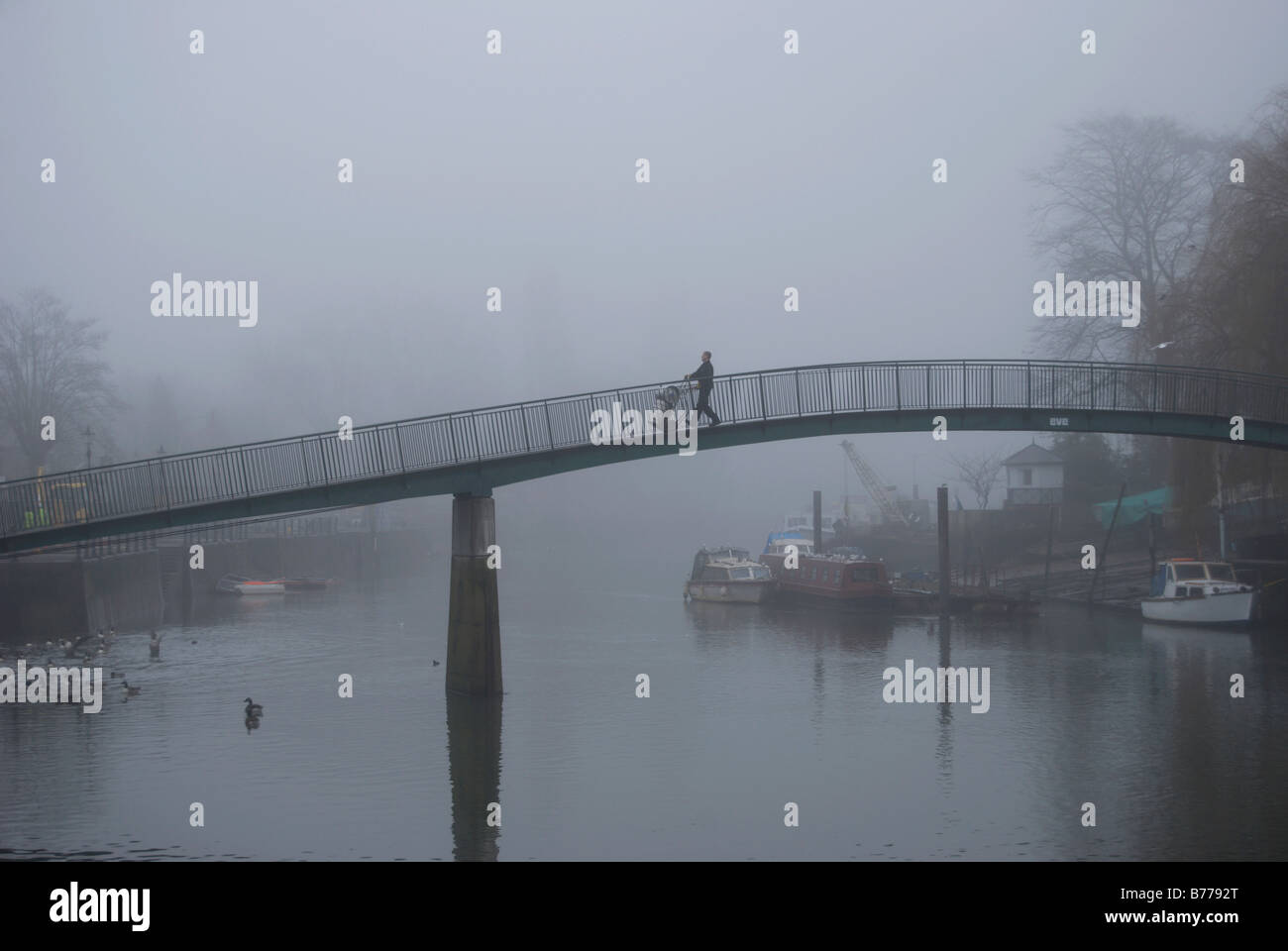 Un uomo si muove di barili di fronte al ponte pedonale sul fiume Tamigi tra eel pie island e il Twickenham su un nebbioso giorno Foto Stock