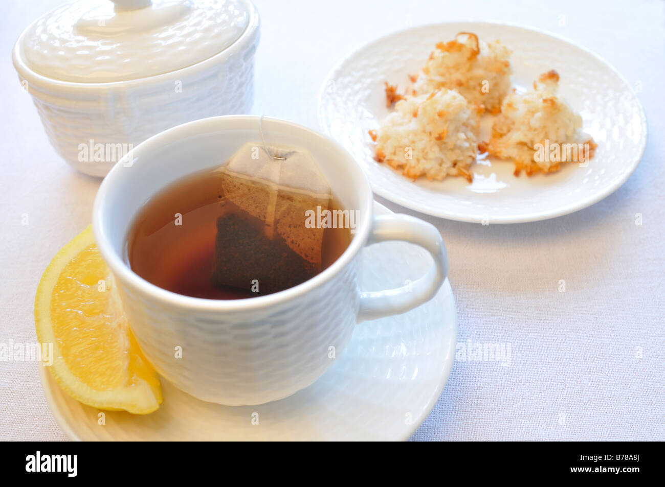Una colazione di impostazione di una tazza di tè nero e limone con una piastra di amaretto cocco biscotti e zucchero su uno sfondo bianco. Foto Stock