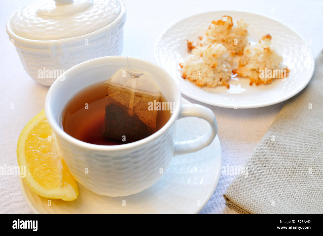 Una tazza di tè nero con una fettina di limone, zuccheriera e piastra di amaretto di cocco i cookie con un tovagliolo su sfondo bianco. Foto Stock