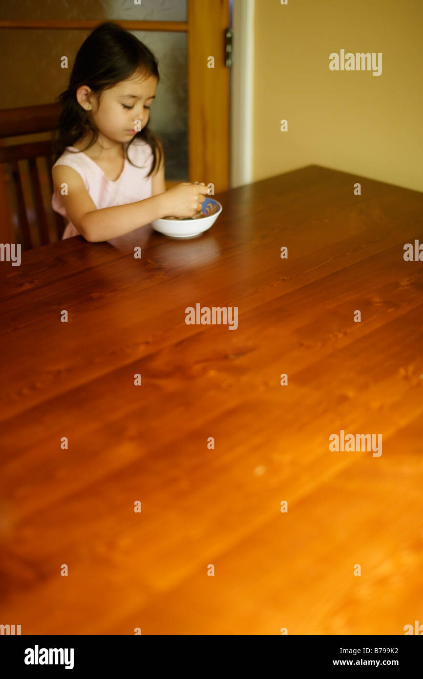 Bambina di cinque anni si siede a un tavolo di legno e si mangia una ciotola di cereale Foto Stock