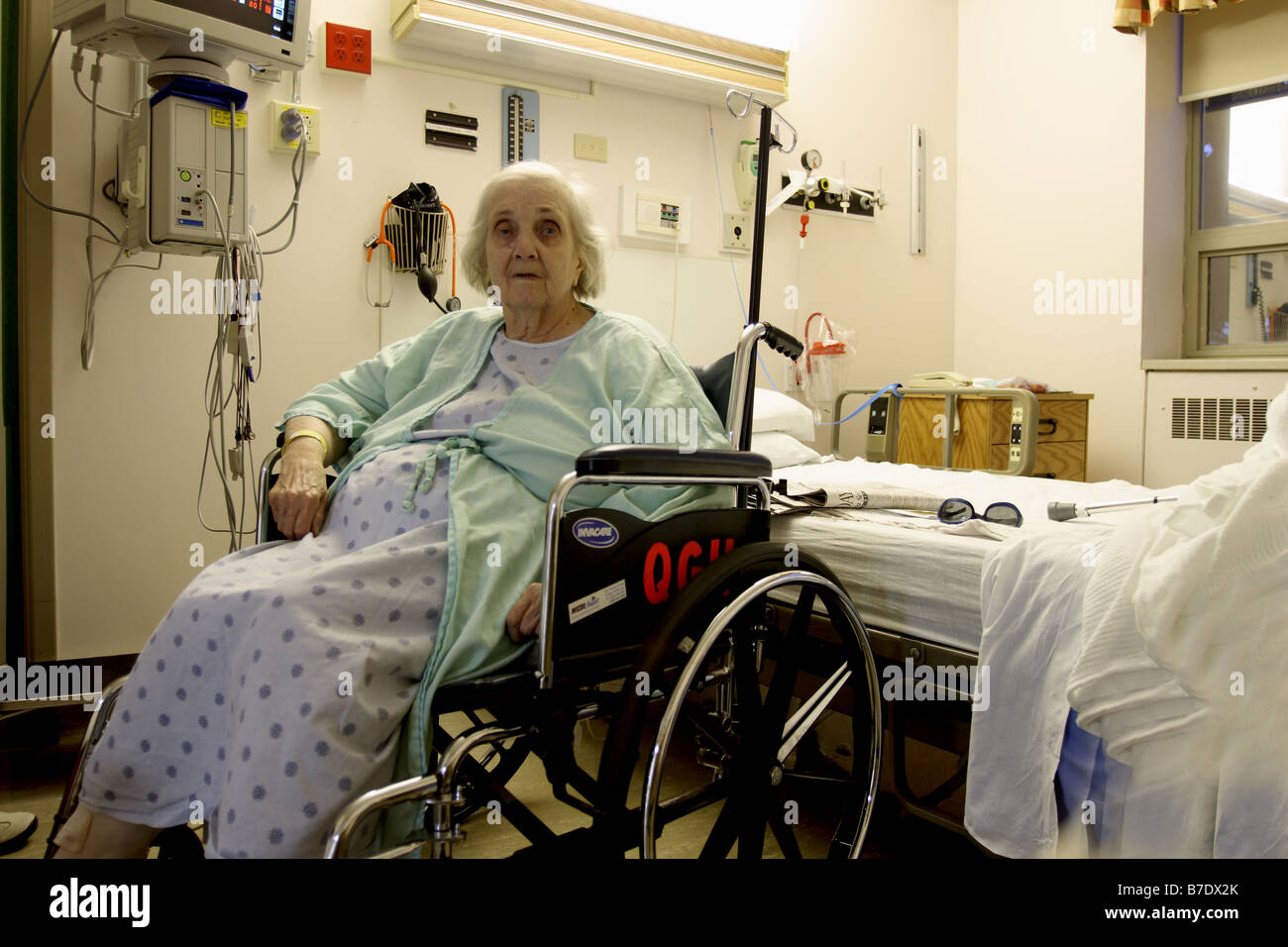 Una donna anziana in un ospedale seduto in una sedia a rotelle nella sua stanza accanto al suo letto Foto Stock