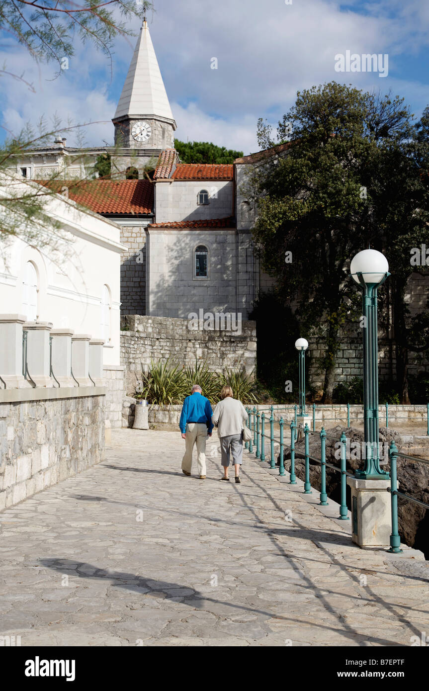 Anziani l uomo e la donna a piedi lungo il lungomare verso la chiesa in Opatija, Croazia Foto Stock