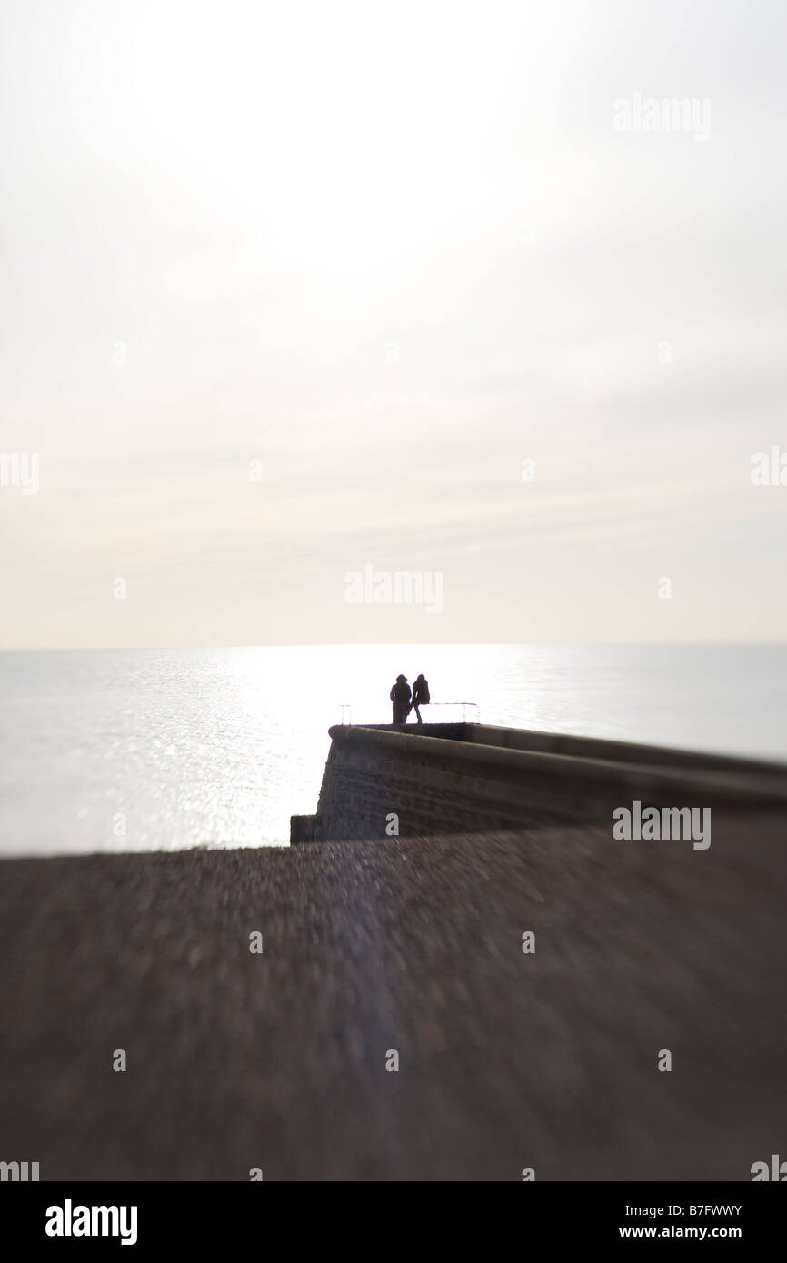 Vista in lontananza due persone sedute al fine di groyne affacciato sul Canale Inglese. Spiaggia di ciottoli in primo piano. Foto Stock