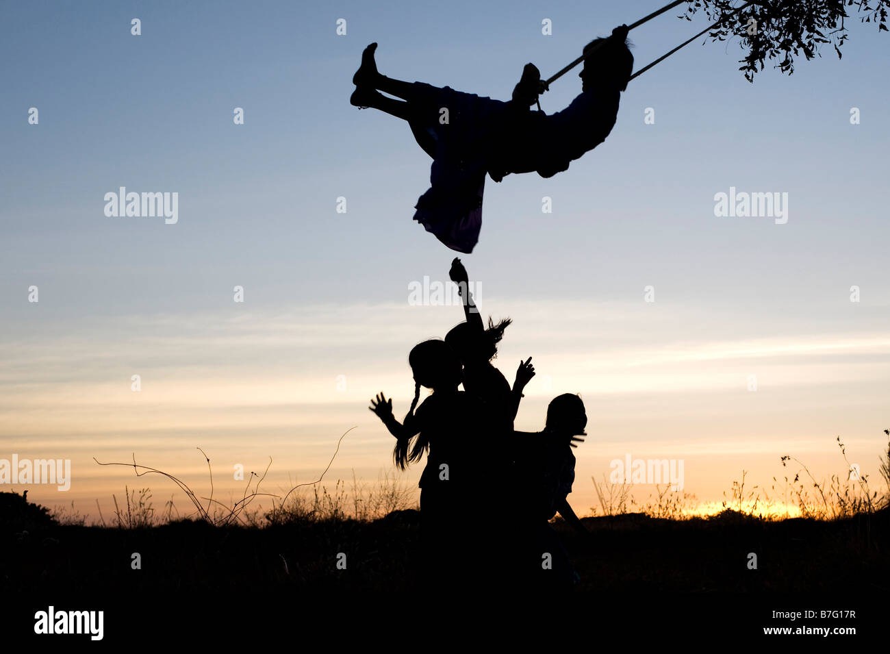 Silhouette di ragazze indiane basculante in una oscillazione in casa nella rurale campagna indiana al tramonto. Andhra Pradesh, India Foto Stock