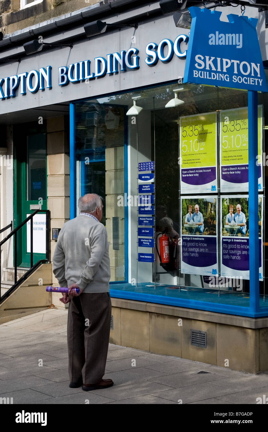 Uomo anziano in piedi fuori dalla filiale della Skipton Building Society che guarda e studia poster (conti, tassi di interesse) - Ilkley, Yorkshire Inghilterra, UK. Foto Stock