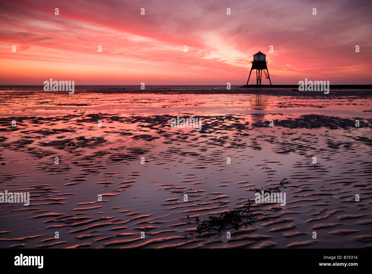 Drammatico tramonto sulla spiaggia con la bassa marea con un faro profilarsi in lontananza. Foto Stock