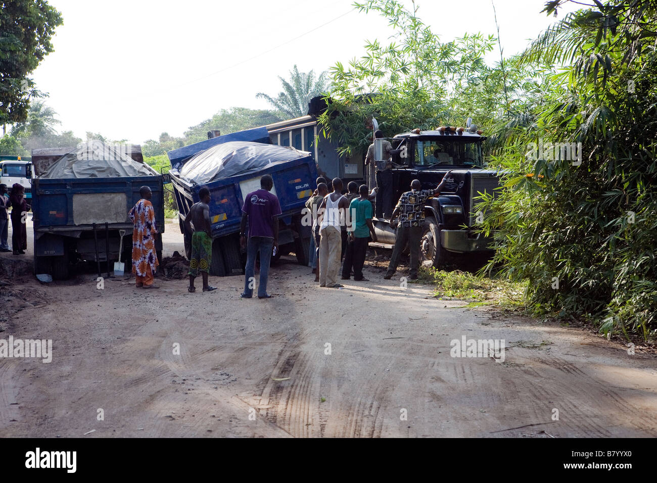Due camion sono bloccati in una trincea fangosa in strada e un terzo autocarro spreme passato spingendo in giù alberi della giungla in Nigeria Foto Stock