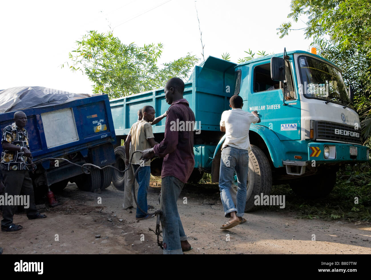 Due camion sono bloccati in una trincea fangosa in strada e un terzo autocarro spreme passato spingendo in giù alberi della giungla in Nigeria Foto Stock
