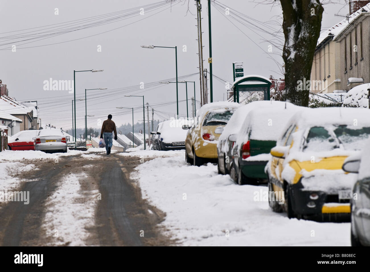 Un uomo a piedi nel centro di una strada coperta di neve veicoli parcheggiati sul lato. Foto Stock