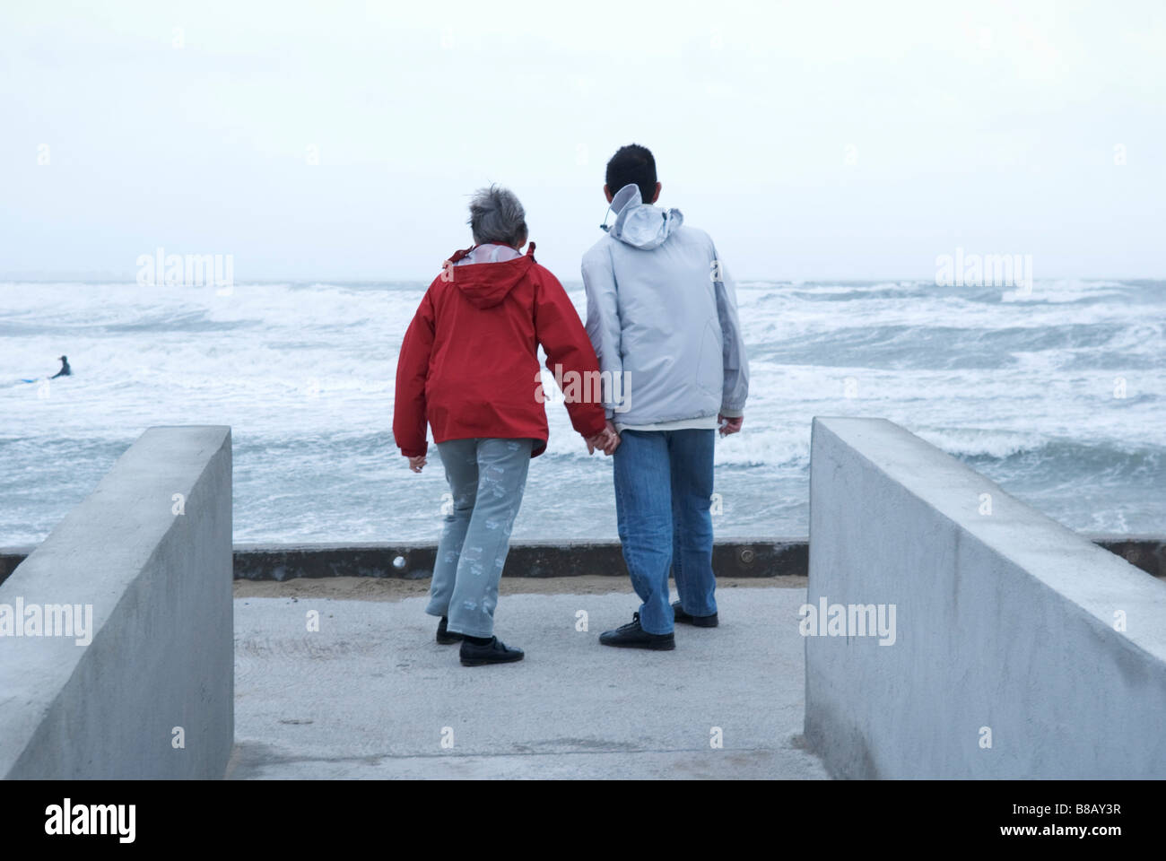 Una vista posteriore di due anziani tenendo le mani e piedi su un lungomare che si affaccia sul mare da un giorno di tempesta Foto Stock