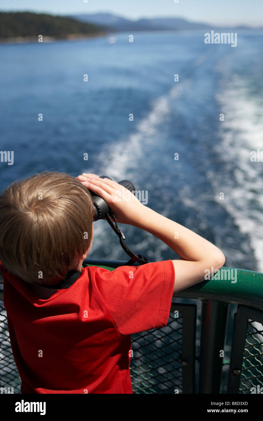 Ragazzo aft deck traghetto cercando in distanza binocolo, traghetto scia visibile Foto Stock