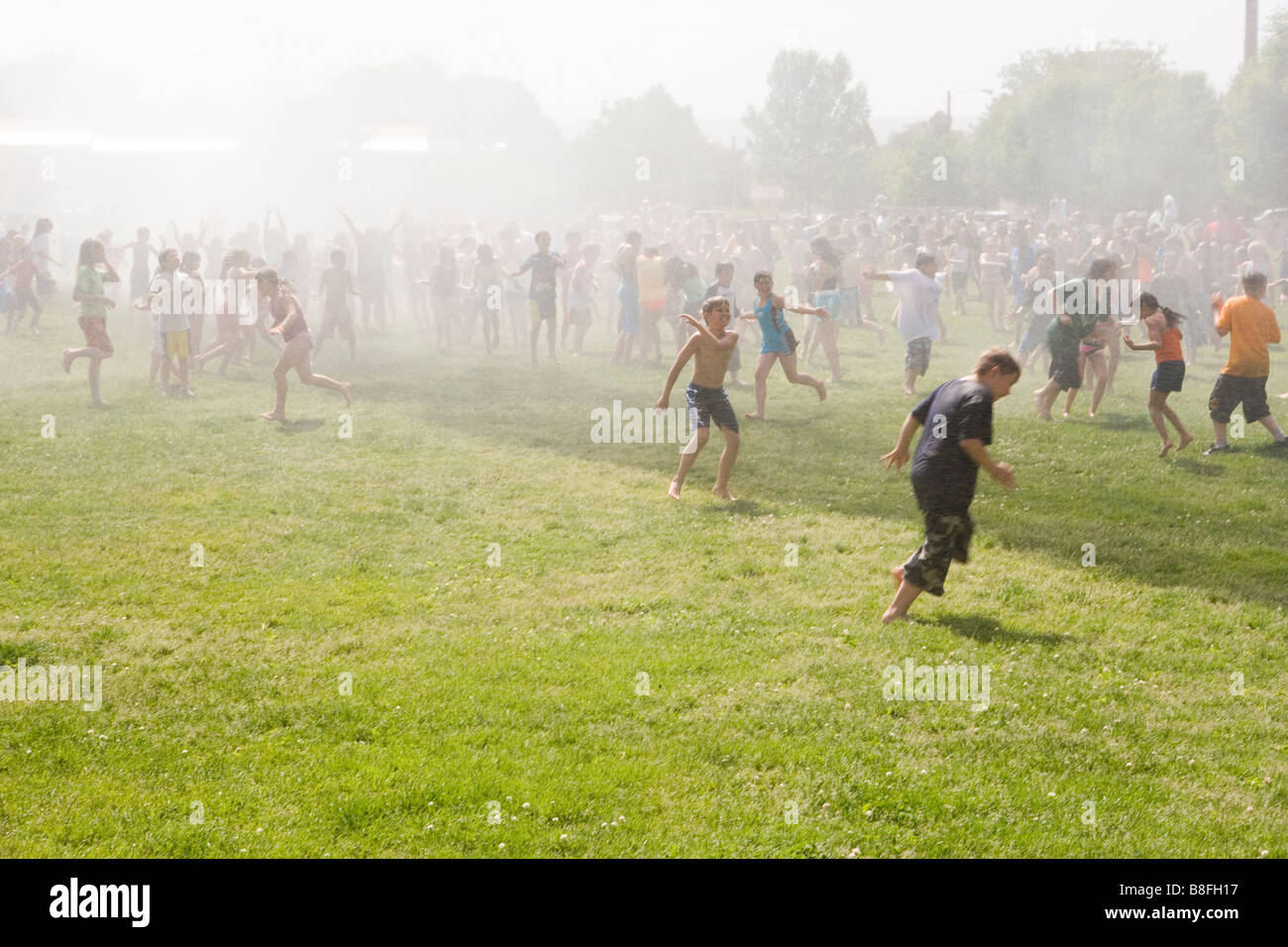 Grande gruppo di età elementari agli studenti in esecuzione con gioia su un campo erboso mentre viene spruzzata con acqua mediante camion fuoco Foto Stock