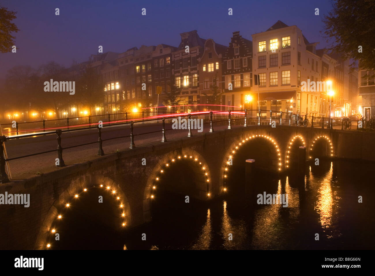 Ponte illuminato riflessa nel canale in una nebbiosa notte a Amsterdam Paesi Bassi Foto Stock